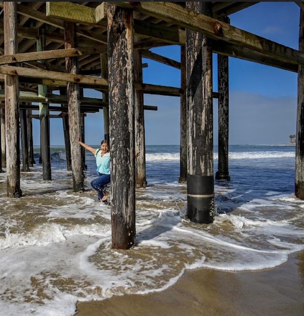 Shantal leaning out from behind a pier piling with her arms open as waves wash in
